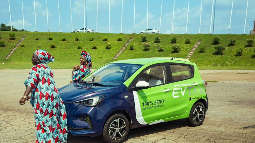 Two women in uniform traditional attires dancing in front of branded electric taxi at the City Gate, Abuja.