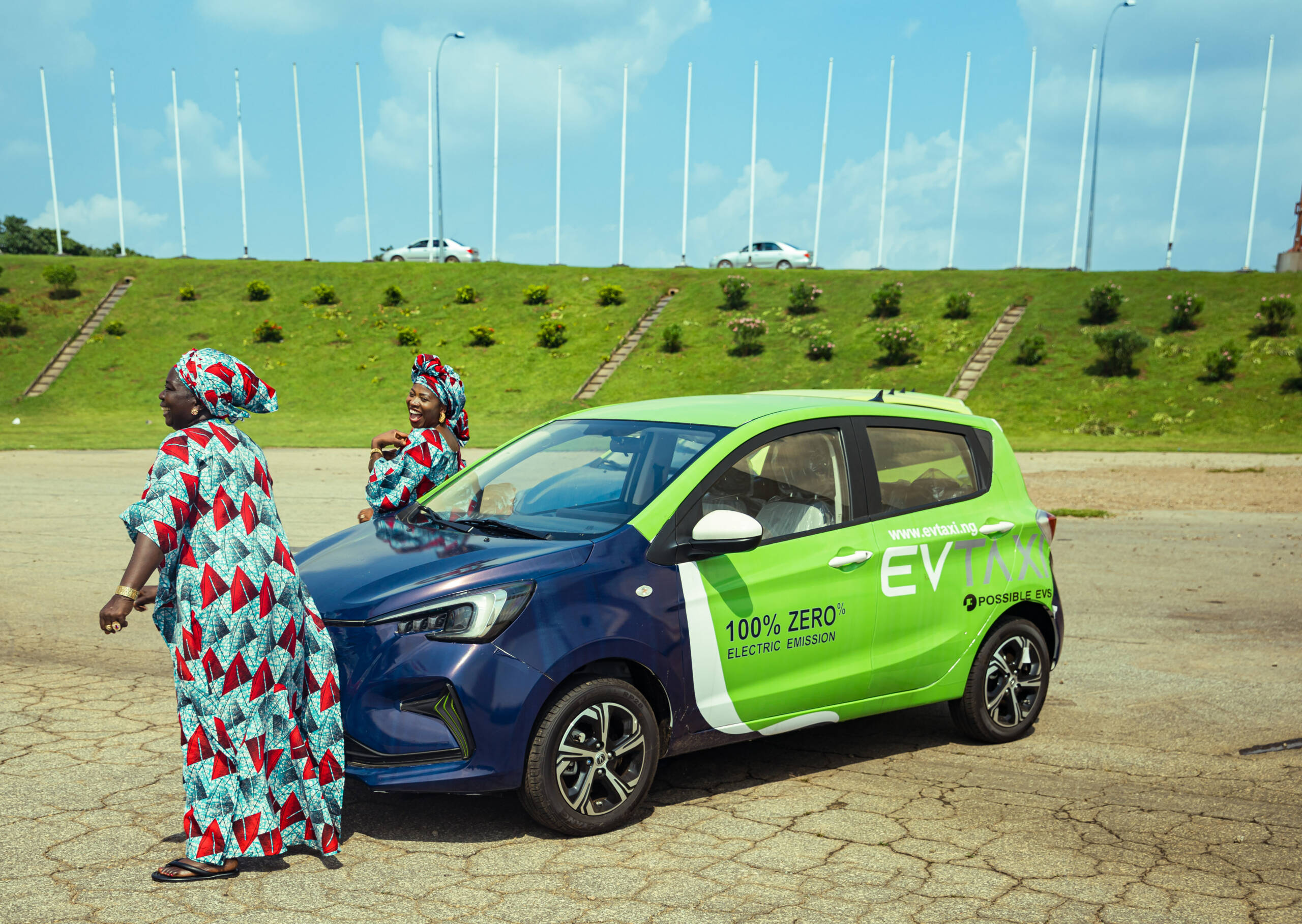Two women in uniform traditional attires dancing in front of branded electric taxi at the City Gate, Abuja.
